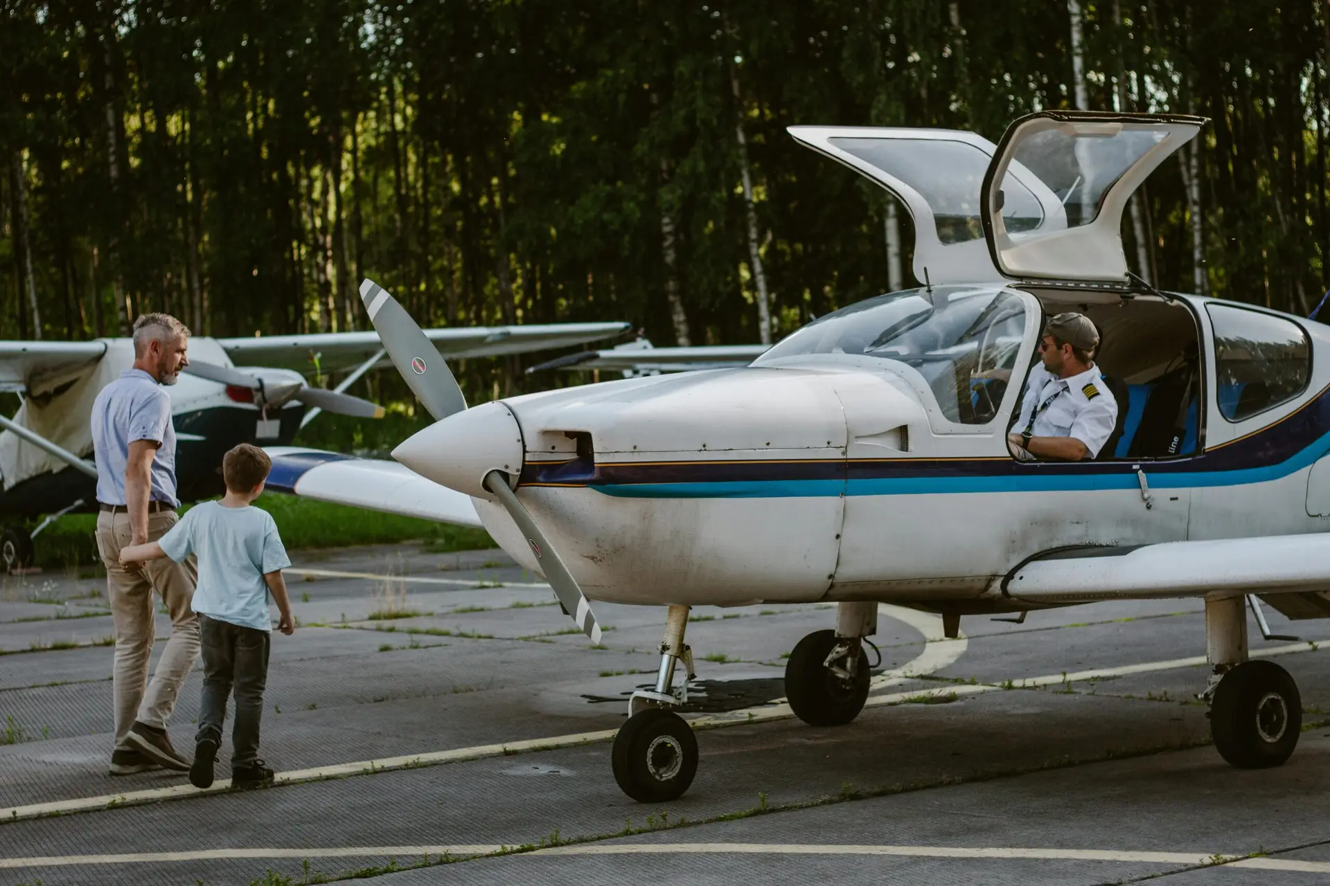 A father and son walking towards a light aircraft with a pilot inside on a sunny day.